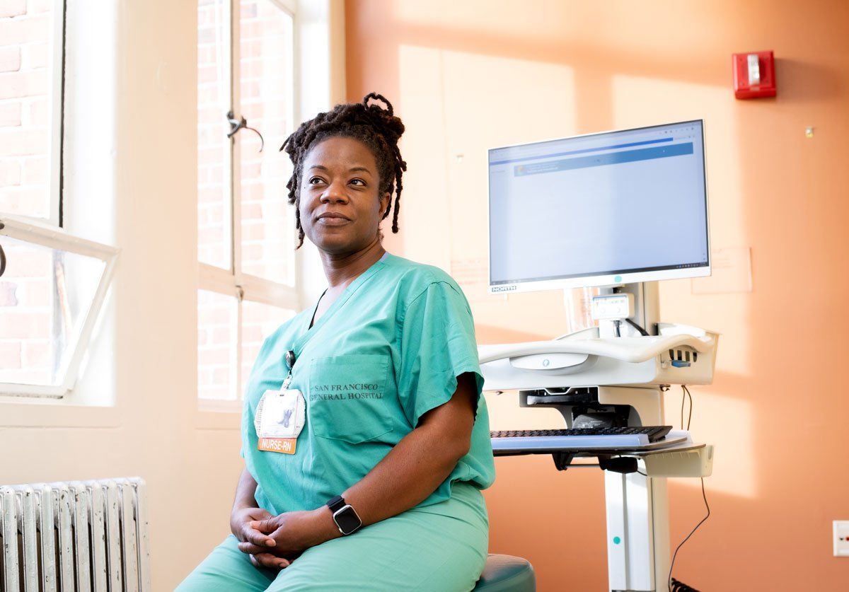 Heneliaka Jones, wearing teal scrubs, sits in front of a computer. Sunlight comes in through the window on a light orange wall