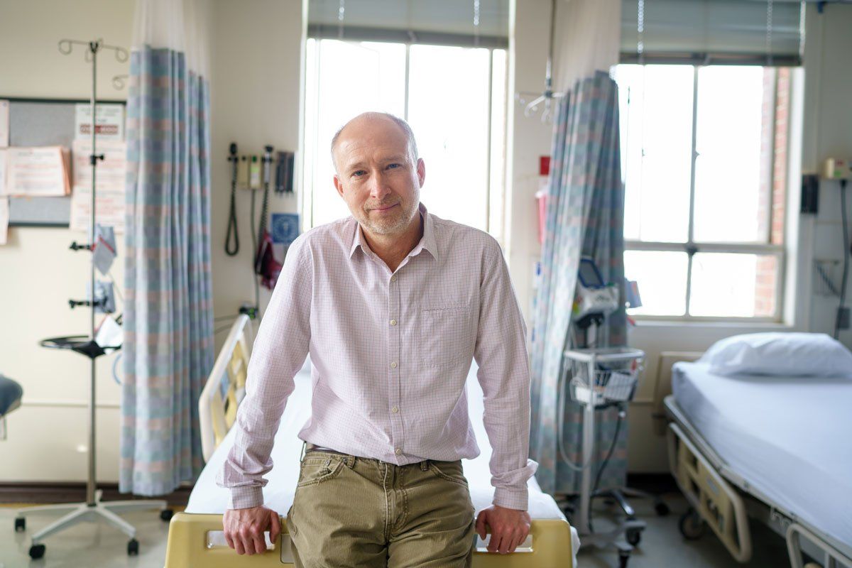 Steve Deeks sits on an examination table in a hospital ward. There are windows and hospital bed curtains in the background