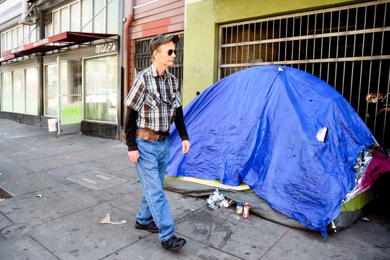 Bill wears a cap and sunglasses as we walks past a blue unhoused tent on a sidewalk.