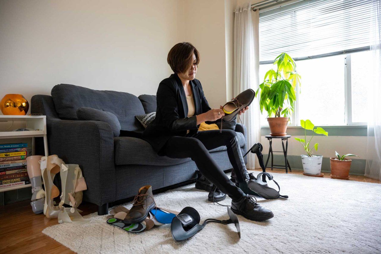 Caro Reyes applies a brace to her shoe in her brightly lit living room. A green plant is in the background.