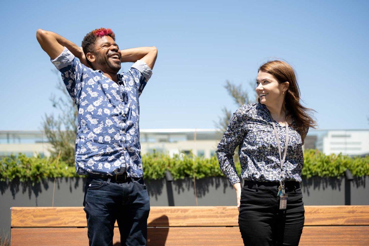 Chase Anderson stands outside under a blue sky and gleefully stretches his arms behind his head while he laughs. His colleague Caitlin stands behind him and smiles as she watches him.