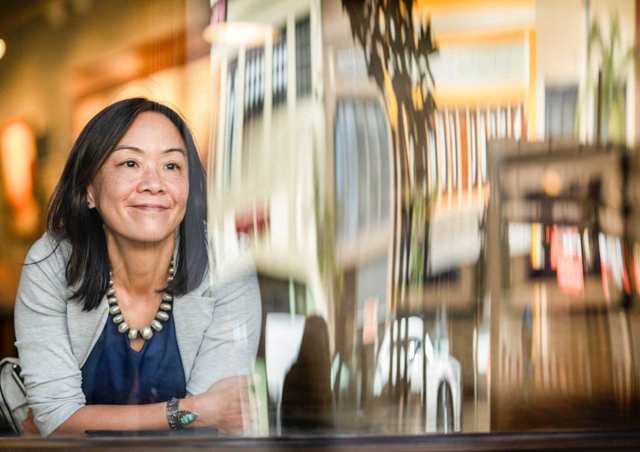 Angela Woon smiles as she looks out a large window, which relfects the buildings on a San Francisco Street.