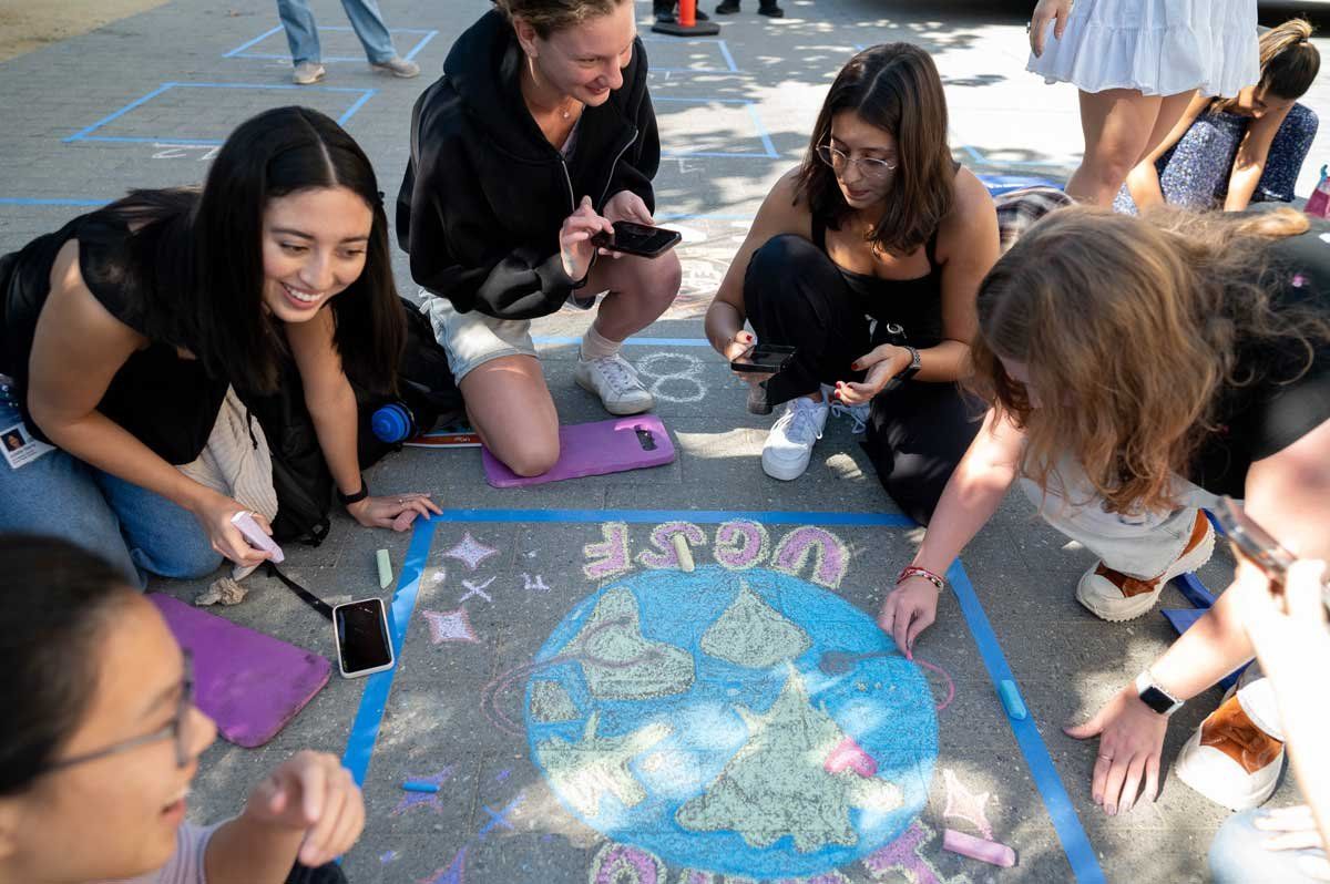 A group masters students smile as they crowd around their chalk drawing of a globe with "UCSF" written below it.