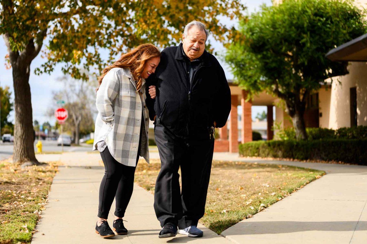 A Hispanic daugther and father duo laugh together as they walk along a sidewalk in Salinas, California. The man's daughter laughs as she leans her head against his shoulder.