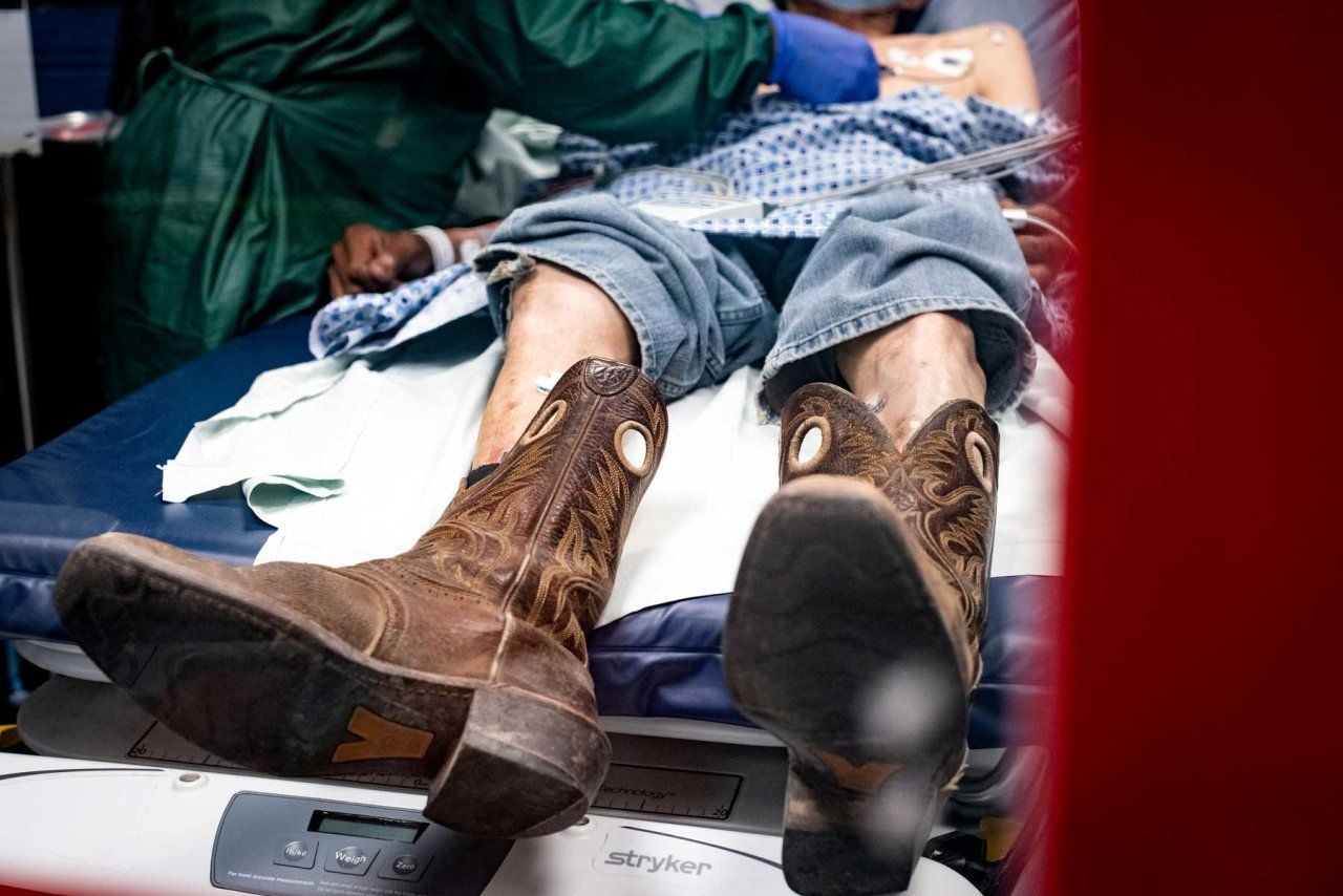 A medical professional gives emergency care to a thin man wearing jeans and cowboy boots underneath a hospital gown. 
