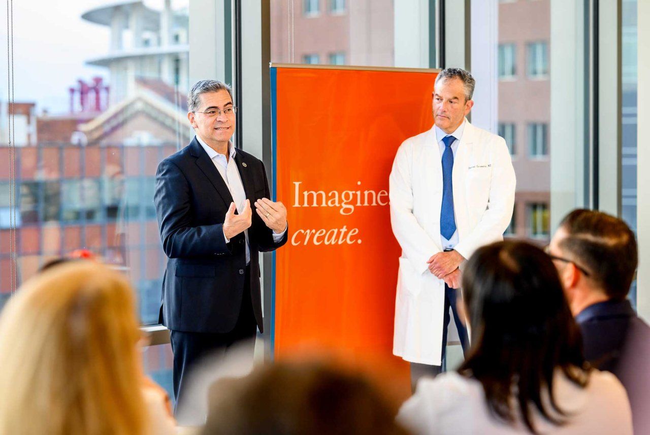 Xavier Becerra (left) speaks to a crowd in a light-filled atrium at Zuckerberg San Francisco General Hostpial. Daniel Grossman stands to his right. In the middle is a large orange banner with the UCSF expression that reads "Imagine. Create."