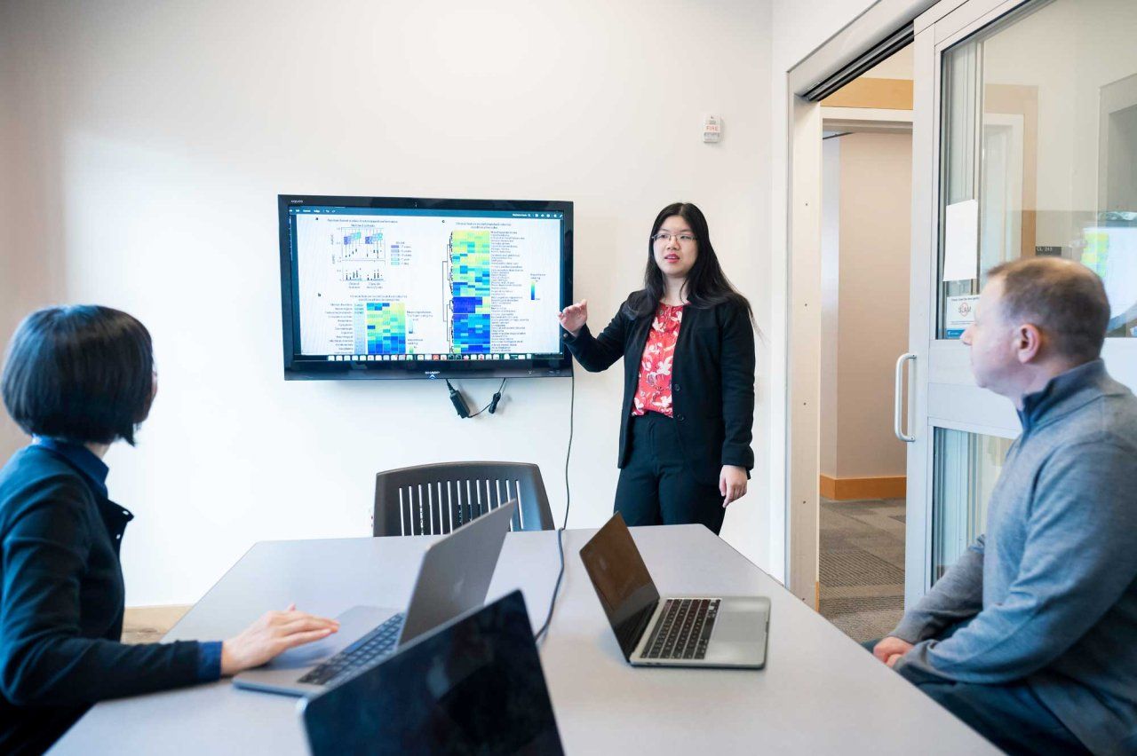 Alice Tang (center) presents scientific research on a TV screen to a man and woman seated at a conference table.