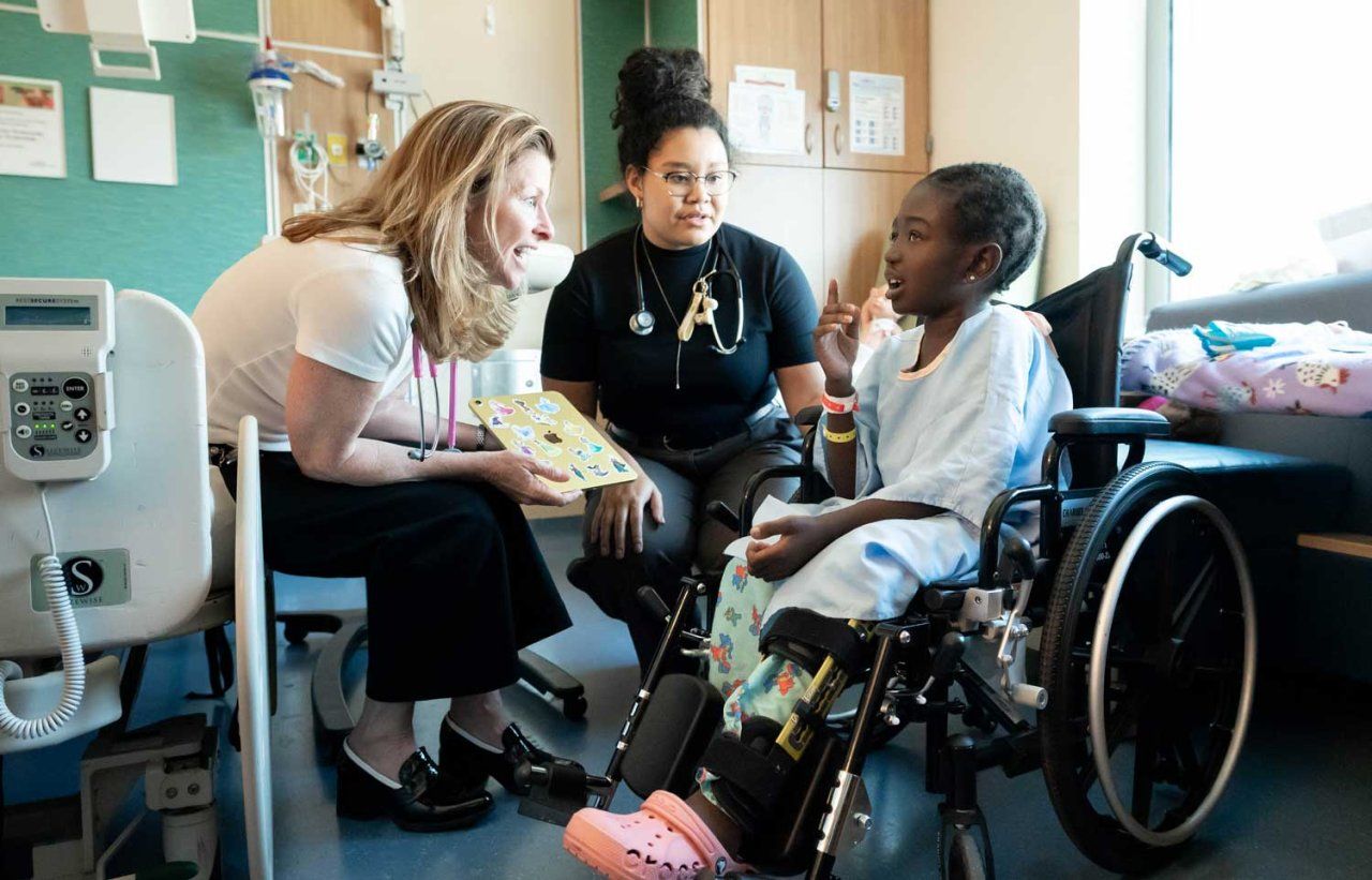 Pediatricians Michele Long and Kathleen Wallace sit with a young patient from Tanzania who sits in a wheelchair.
