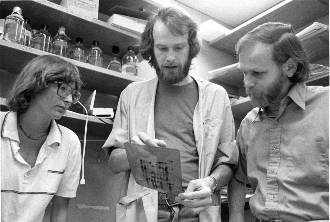 A black and white photo of Michael Bishop in a lab with colleagues in 1989.