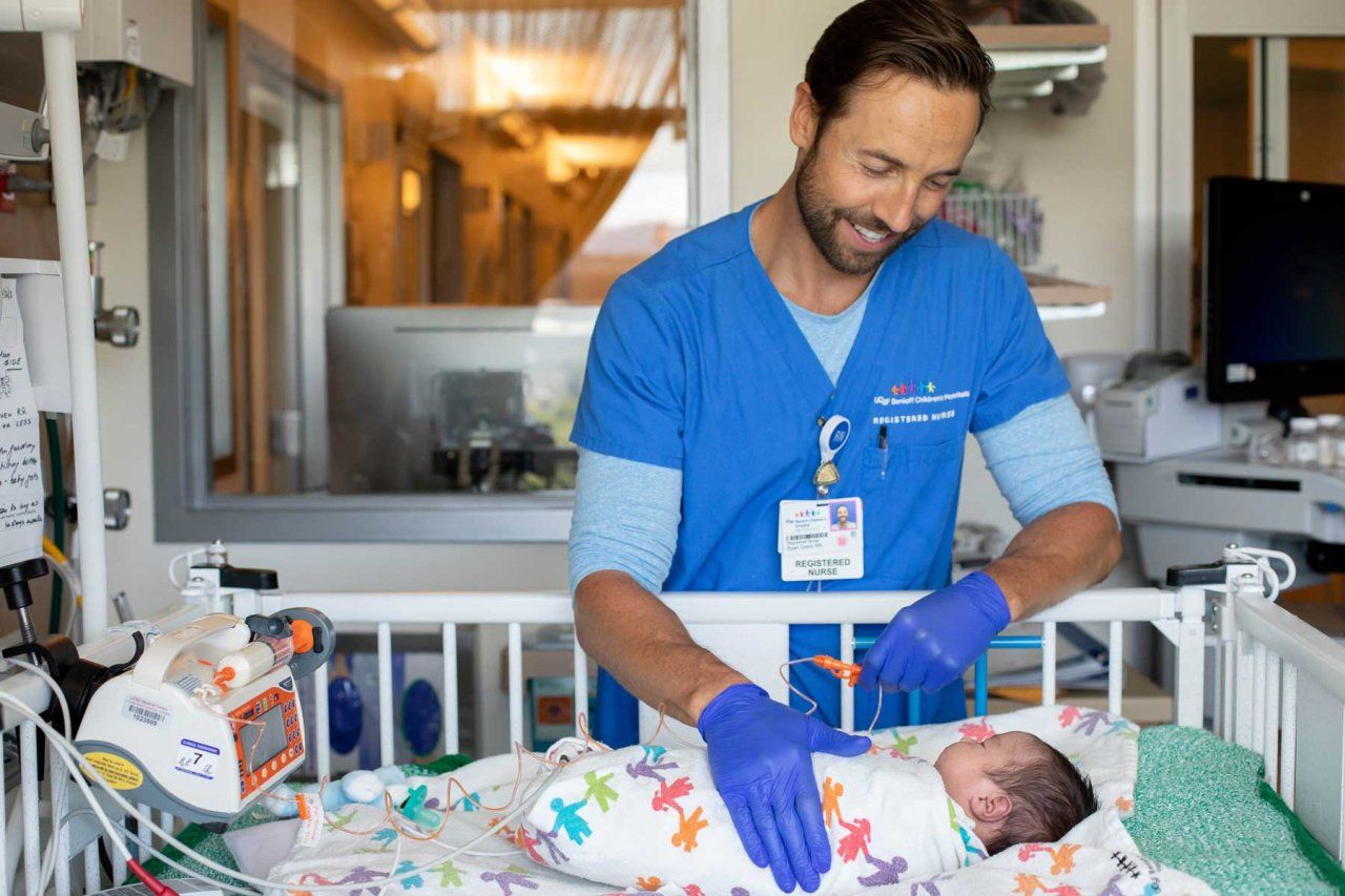 A registered nurse smiles as he tends to a baby in a pediatric intensive care room.