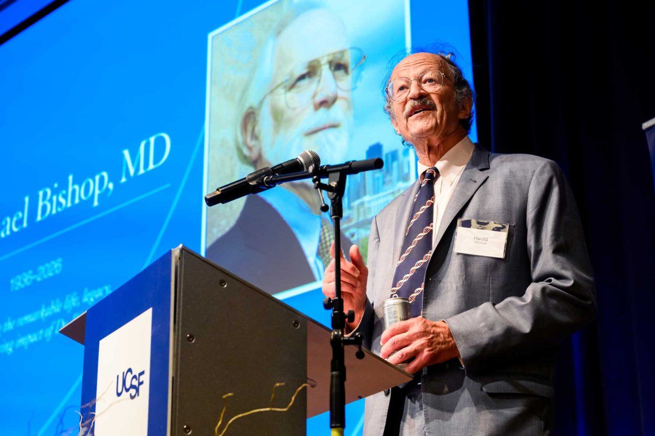 Harold Varmus stands at a podium with a screen behind him showing a photo of J. Michael Bishop.