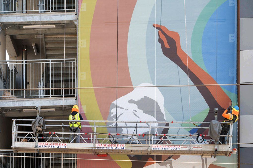 A mural of a boy flying a kite being installed by two workers on scaffolds along the side of a parking garage wall.