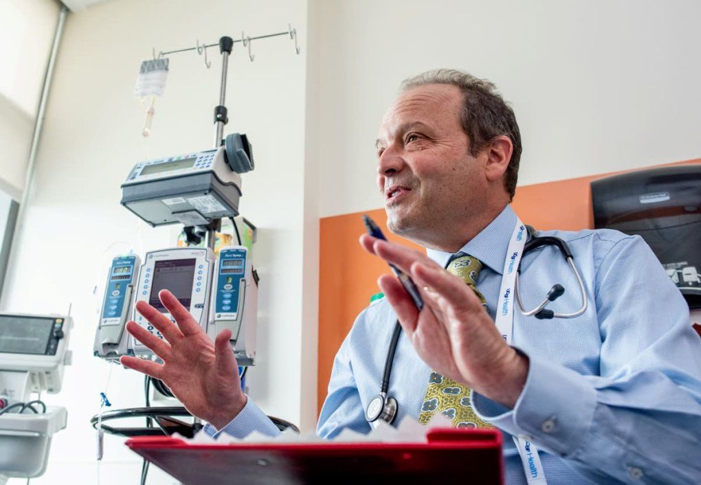 A male doctor wearing a stethoscope uses manual gestures while speaking. Medical equipment is in the background.