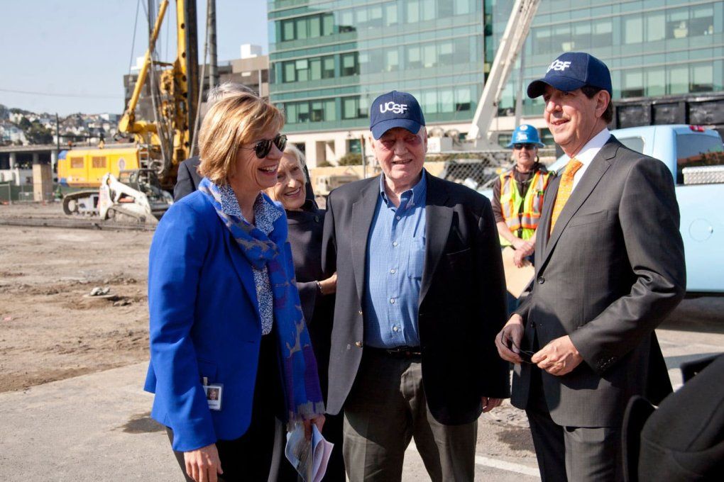 A woman with a blue blazer and two men wearing blazers and blue hats that read "UCSF" stand at a construction site. A man in reflective gear stands behind them.