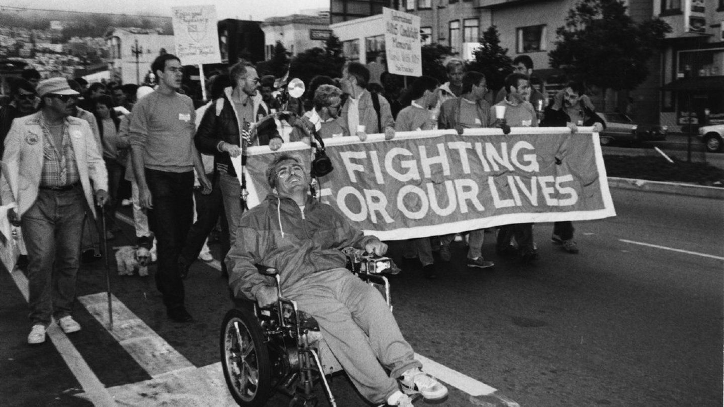 Men carrying a banner that reads Fighting For Our Lives march along San Francisco city streets with a man in a wheelchair in the lead