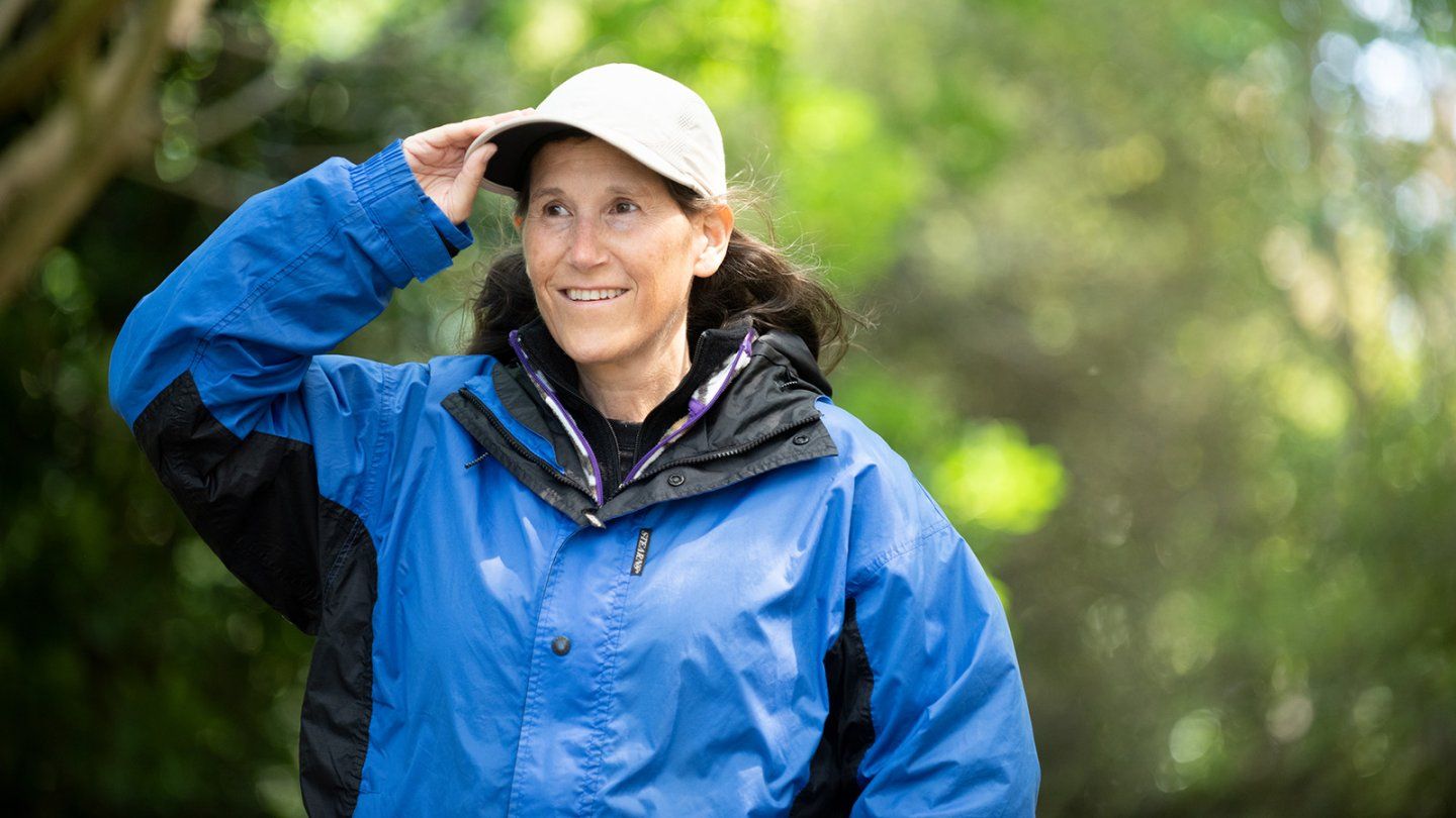 Cheryl Broyles on a hike smiles and lifts her hand to adjust her cap