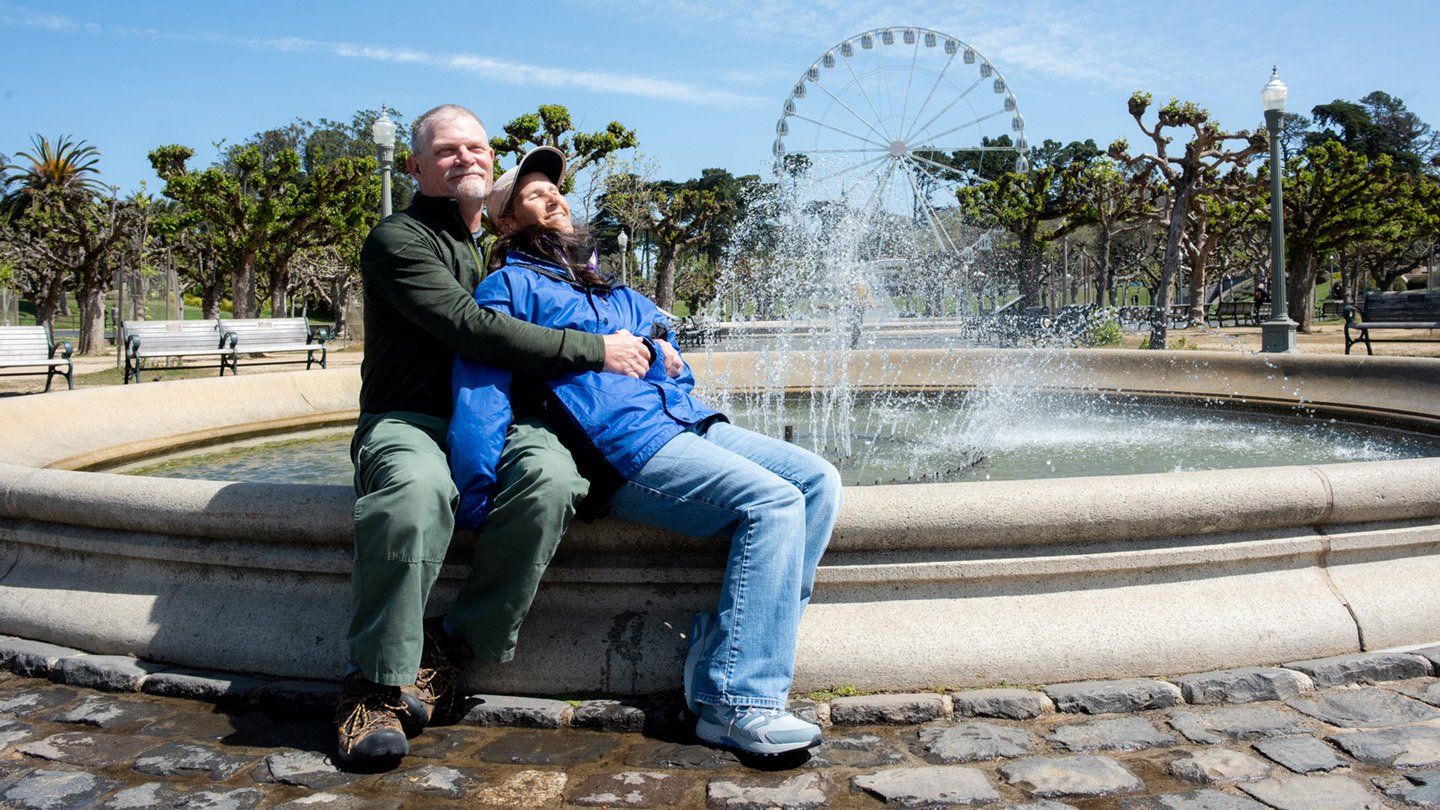 Cheryl and her partner embrace while sitting at the edge of a fountain in Golden Gate Park