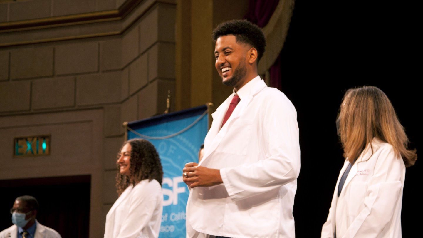 New doctorate students with white coats stand and smile onstage at a white coat ceremony