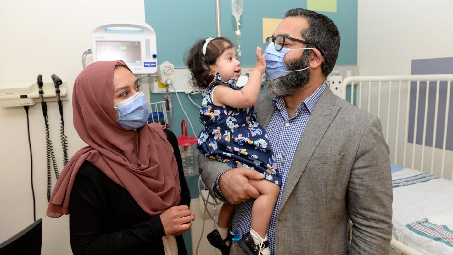 Sobia Qureshi, wearing a mask, smiles as baby Ayla grabs her father Zahid's mask while he holds her