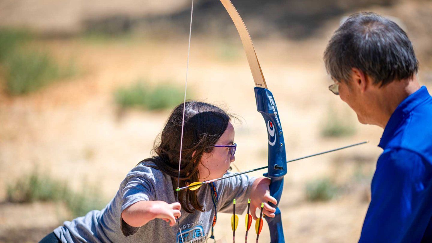 A girl with limb differences practices archery as a counselor smiles and watches on.
