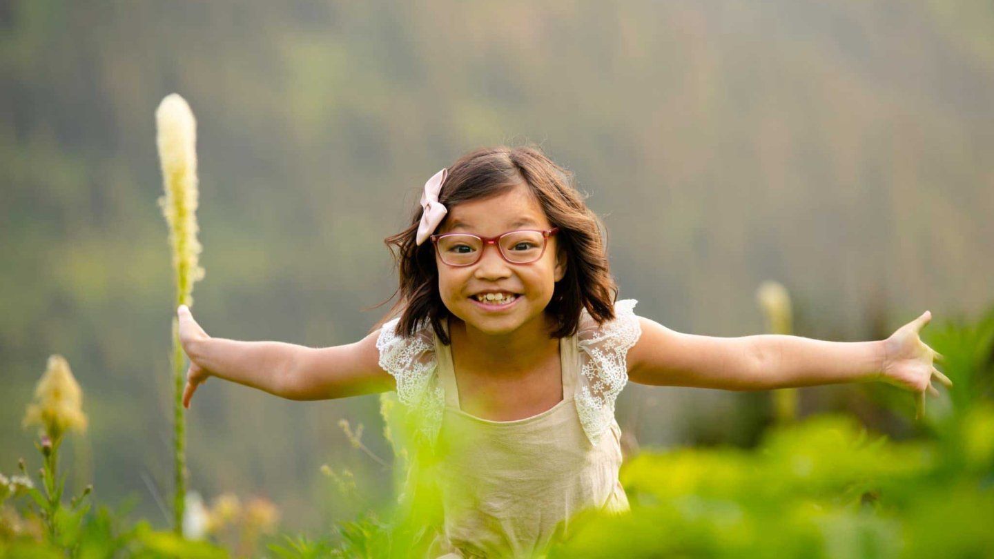 A little girl of Chinese descent smiles with her arms outstretched as she stands in a grassy field.