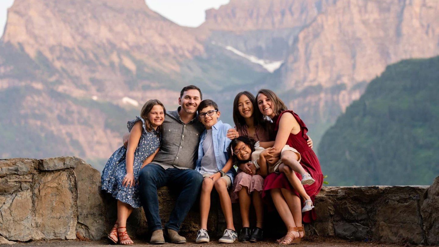 Clint and Alissa Finlayson pose with their three daughters and son for a family portrait in front of a mountanous background.
