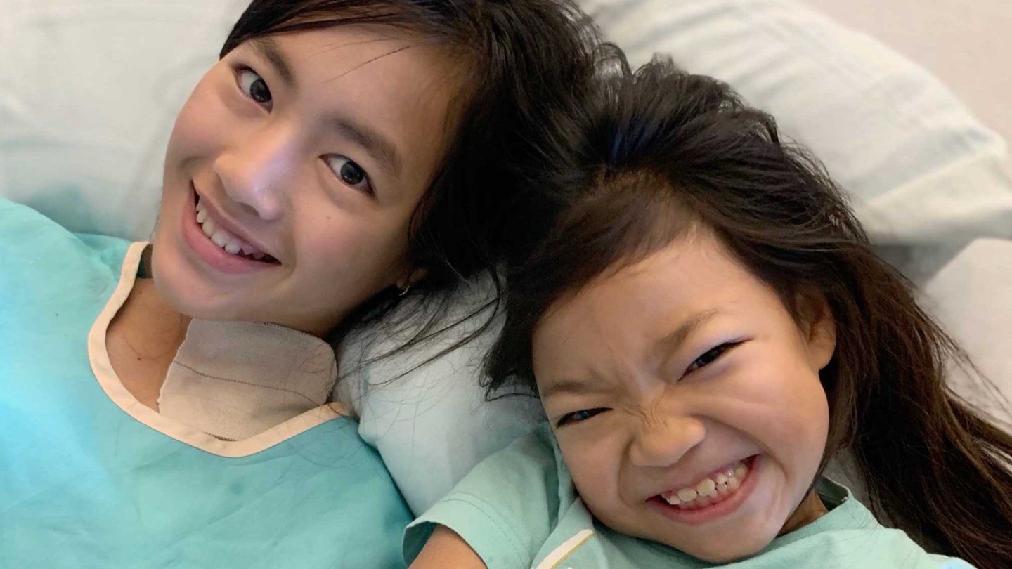 Two young girls, who were adopted from China by an American family, smile as they lay together on a hostpial bed wearing hostpial gowns.