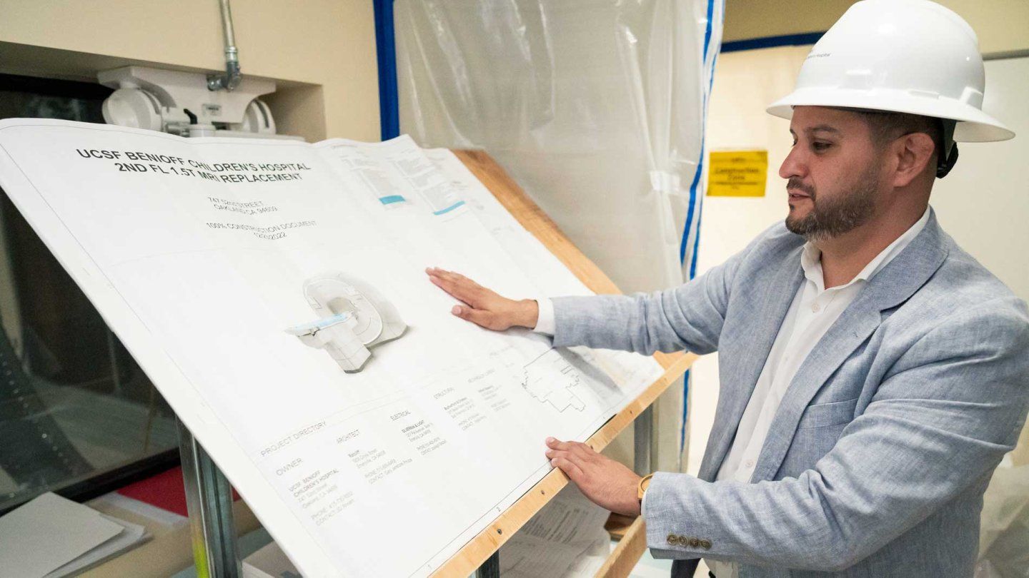 Construction project manager Michael Valero wears a white hard hat while looking at blueprints of an MRI.