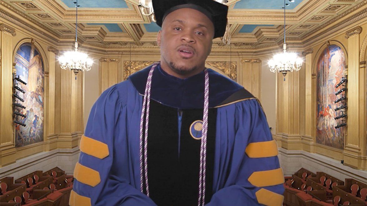 A man in a cap and gown stands before an empty auditorium on Zoom