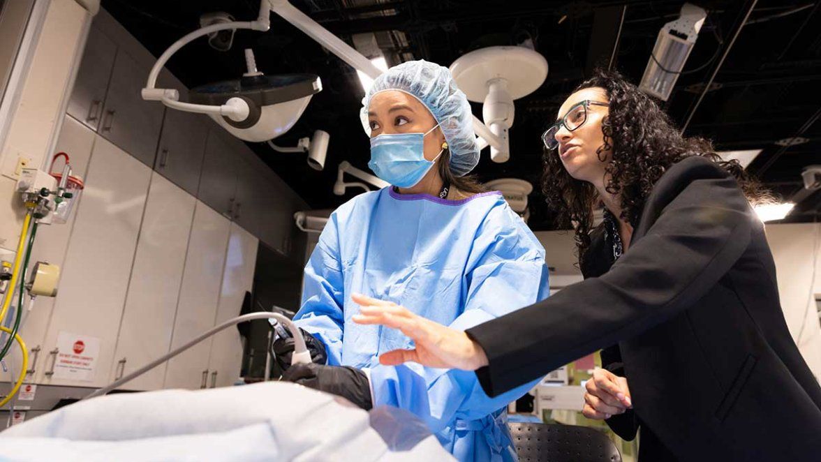 Clinician in surgical attire practices a medical procedure with an instructor in a simulation room.