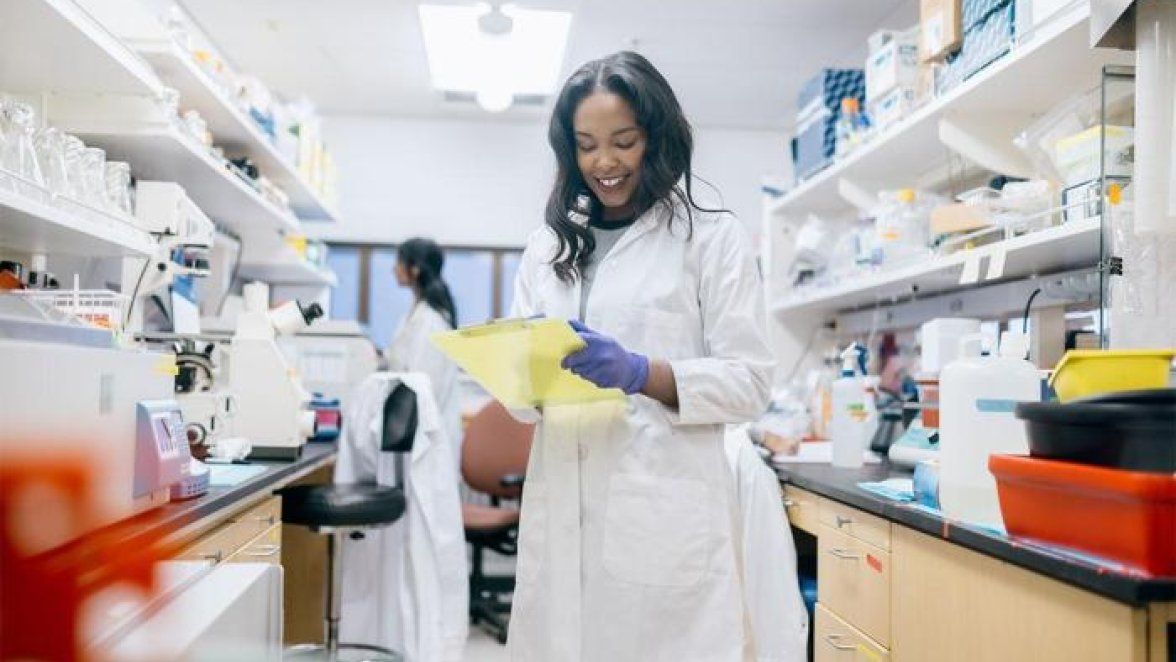 A Black female pharmacist wearing a white coat works in a pharmacy.