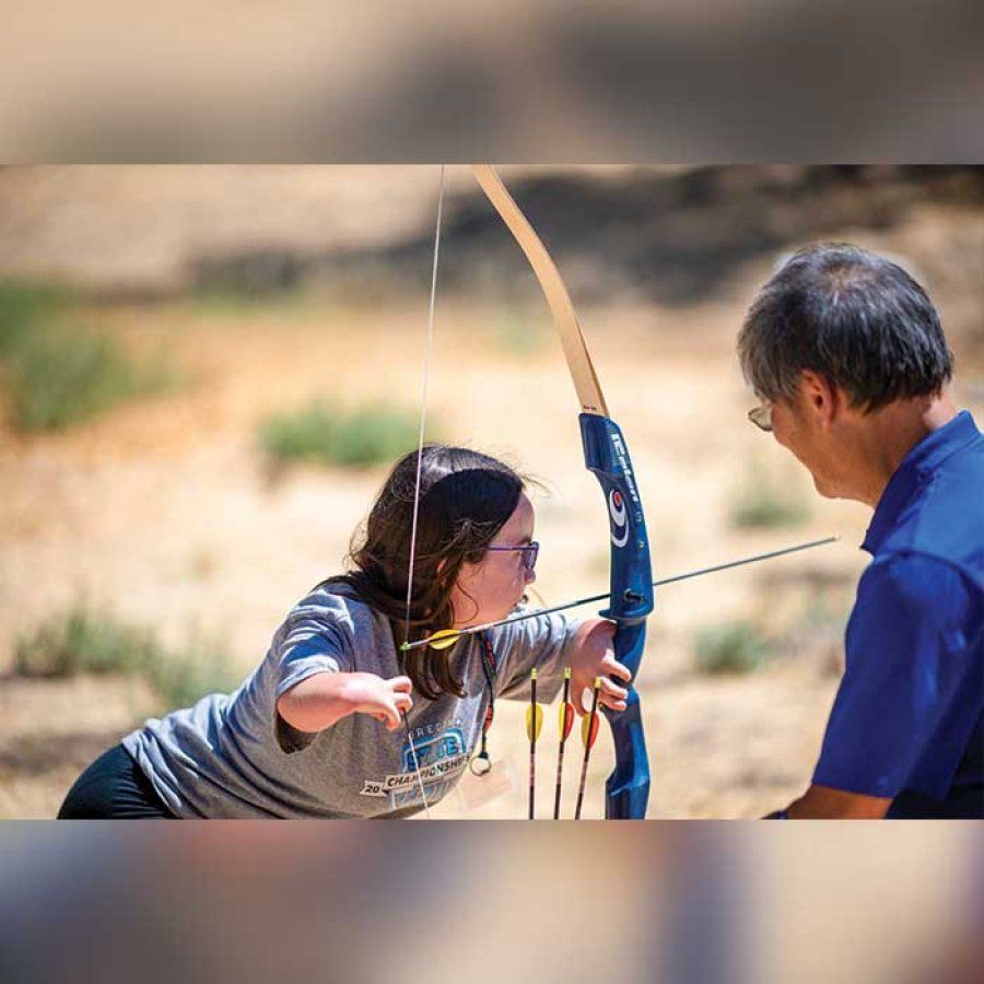 A girl with limb differences practices archery as a counselor smiles and watches on.