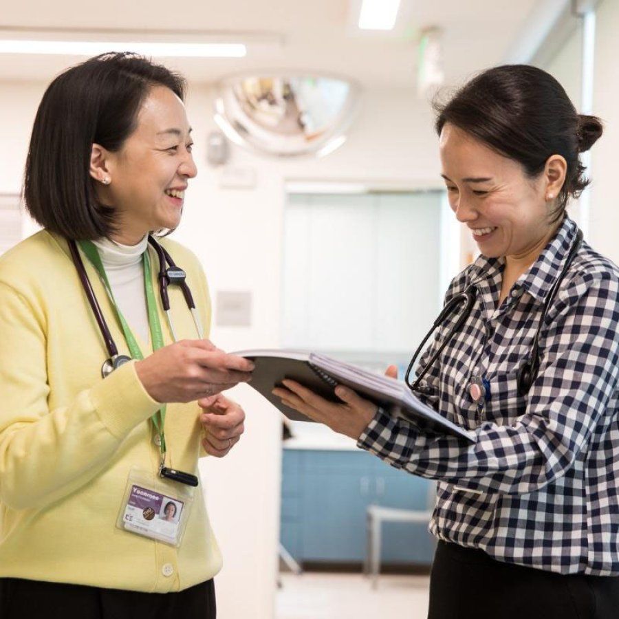 Two women smile and talk, one wearing a stethoscope and the other holding a book.