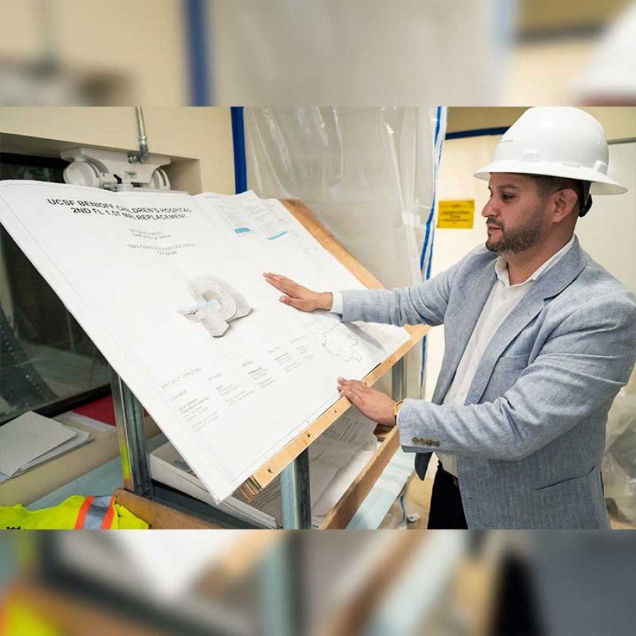 Construction project manager Michael Valero wears a white hard hat while looking at blueprints of an MRI.