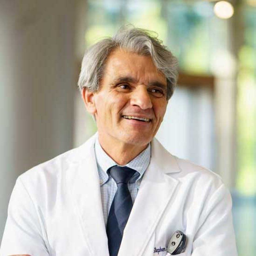 Stephen Hauser smiles as he stands in a light-filled atrium and wears a white doctor's jacket.