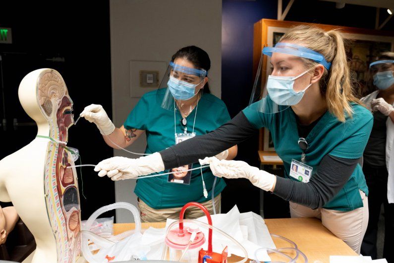 Two students use a plastic tube on an anatomical figure while wearing masks