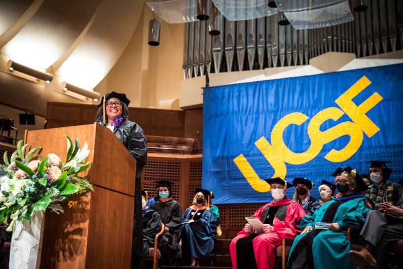 A school of nursing graduate stands at a podium with faculty sitting behind. A banner reading "UCFS" is in the background