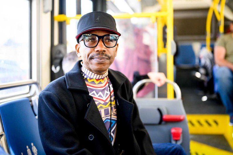 An African American smiles as he rides a bus. He wears a cap and glasses.
