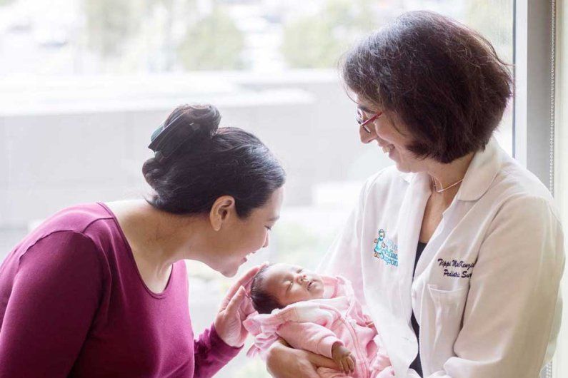 Neonatal surgeon Tippi Mackenzie holds a newborn baby as her mother smiles and holds the baby's head.