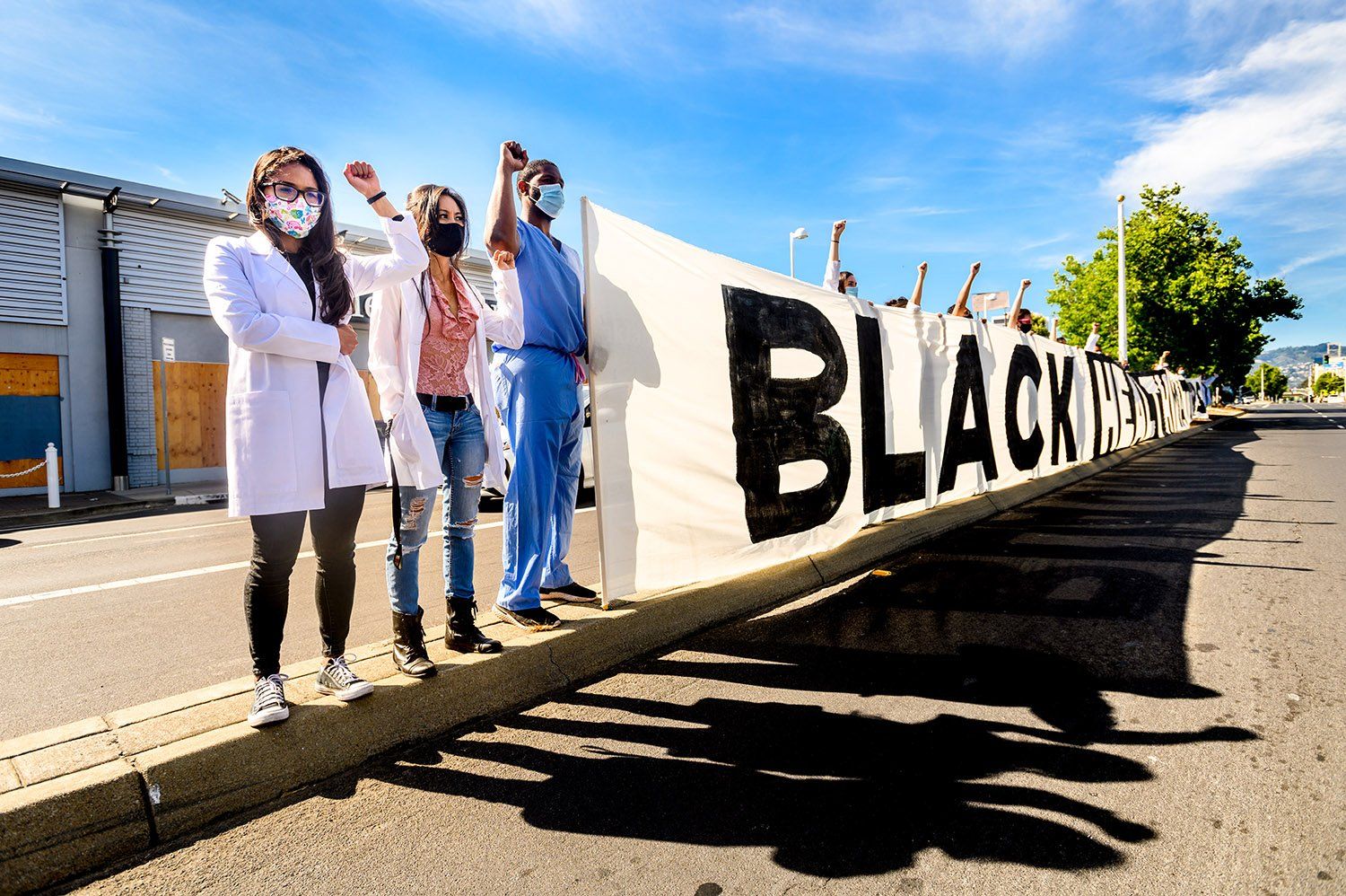 Students stand on a curb in protest wearing masks and holding their fists in the air beside a giant black health matters sign