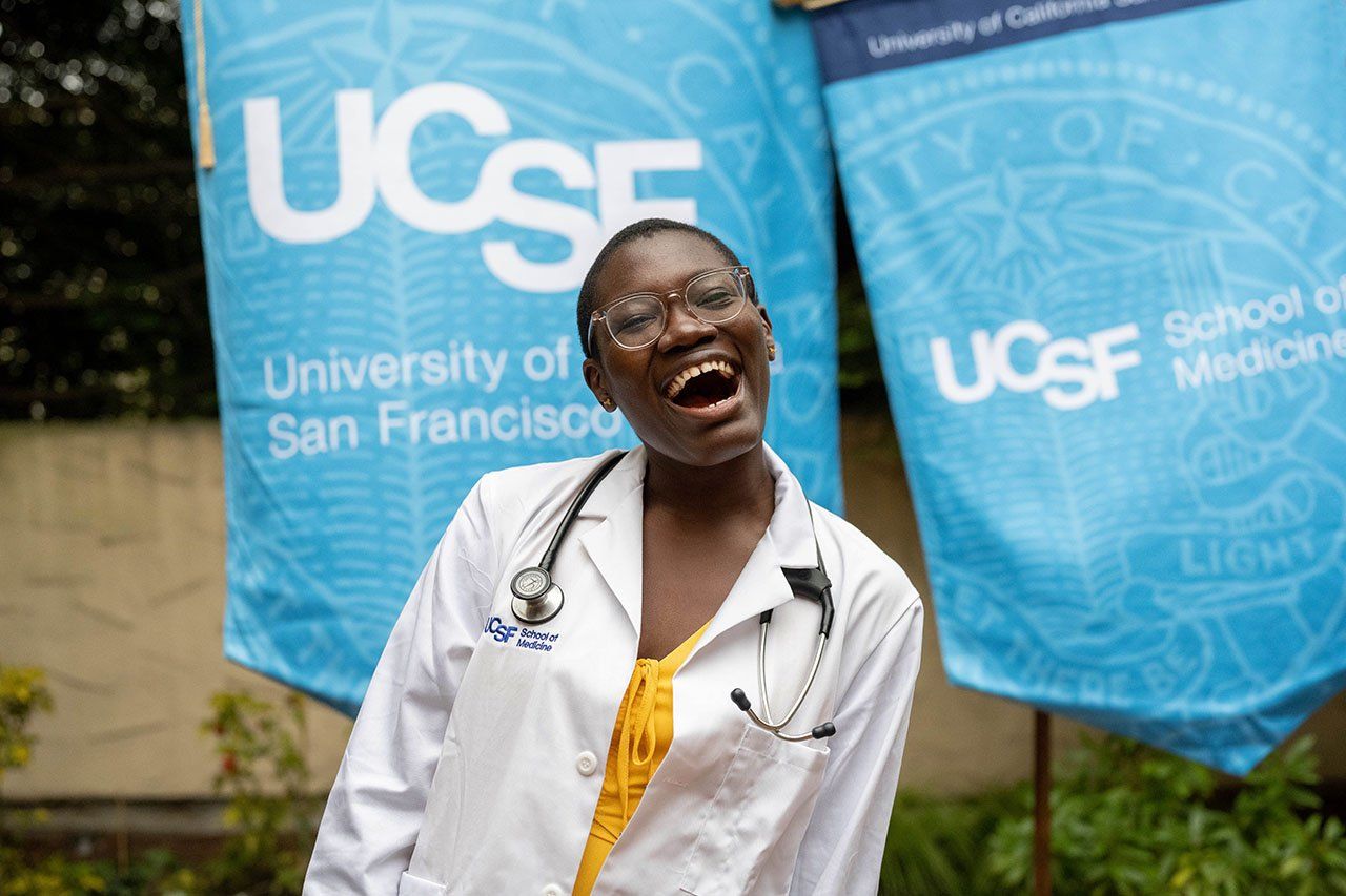 A student with a big smile poses in her new white coat against a background of UCSF banners