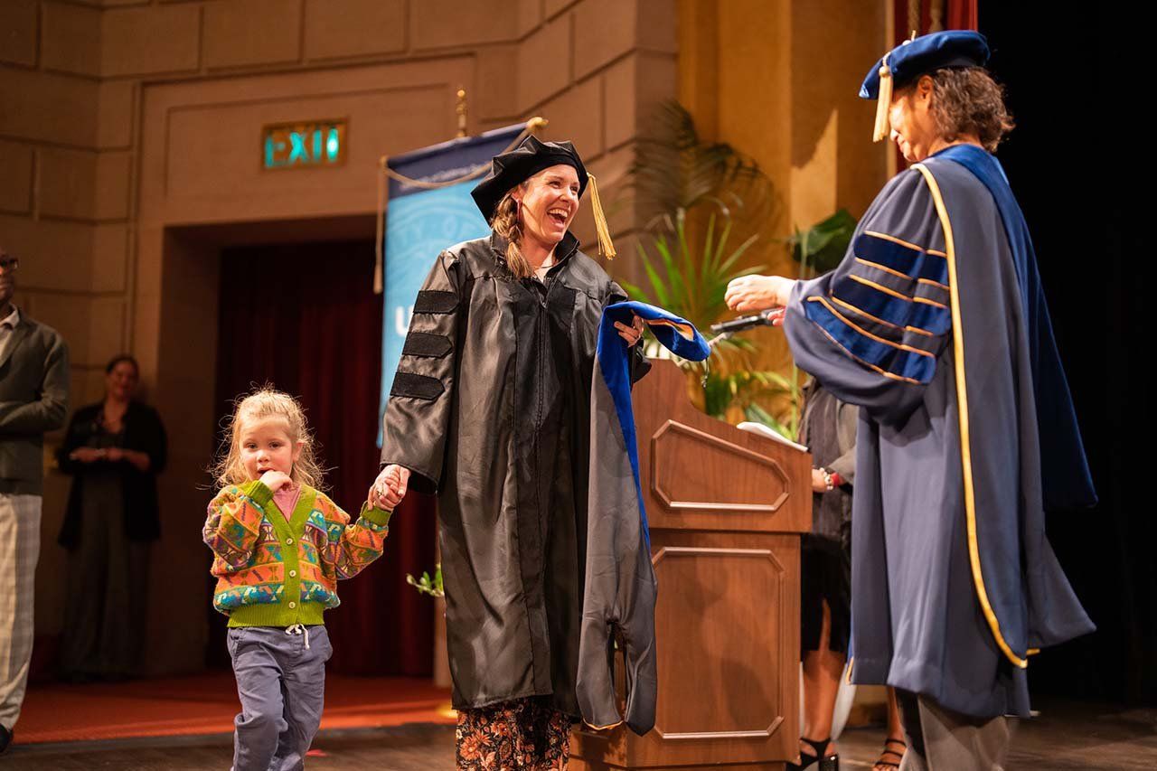 A new graduate grins wildly as she walks across the stage holding the hand of her young child.