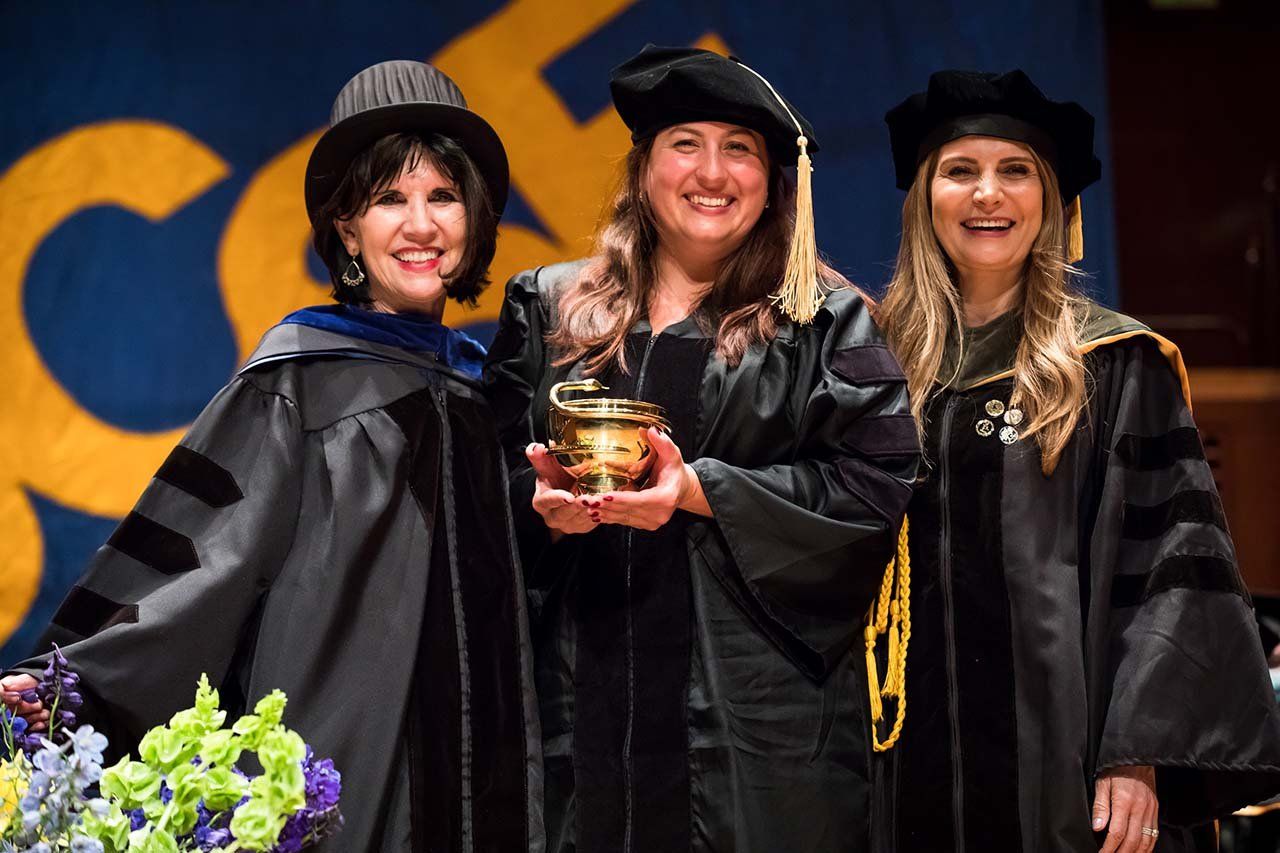 Three people pose on stage before a UCSF banner while the middle holds a golden cup.