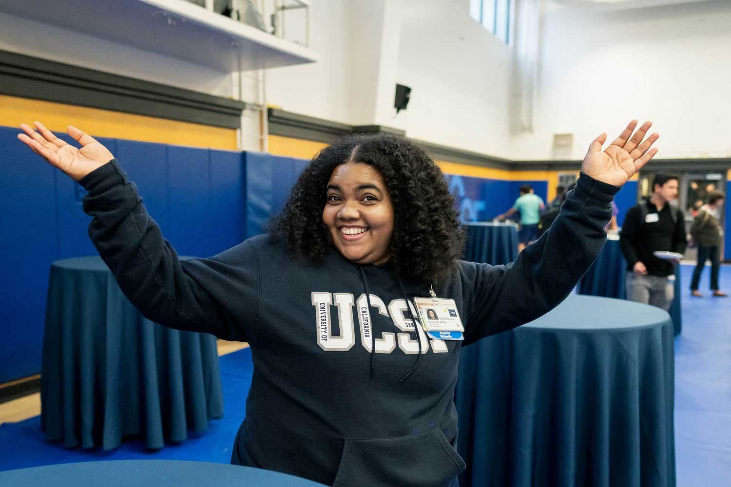 A nursing student wearing a sweatshirt with the UCSF logo smiles and stretches out her hands to pose for a photo.