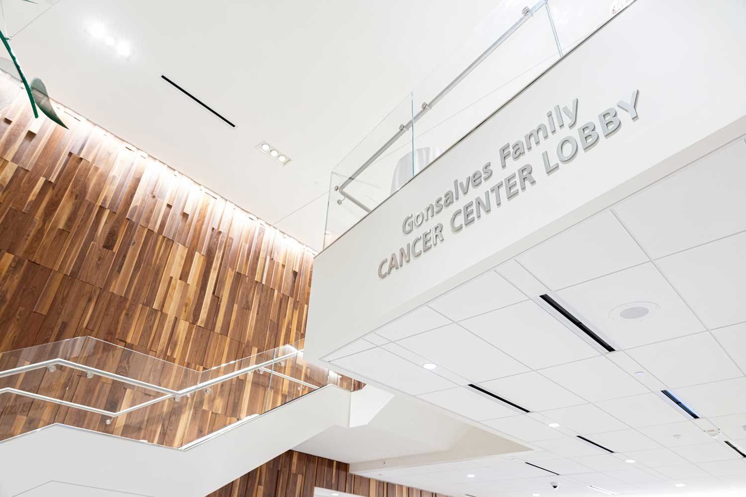 The interior of the Behring Pavilion, showing the Gonsalves Family Cancer Center Lobby, which has a modern wood-paneled wall, and white stairs with glass panes.