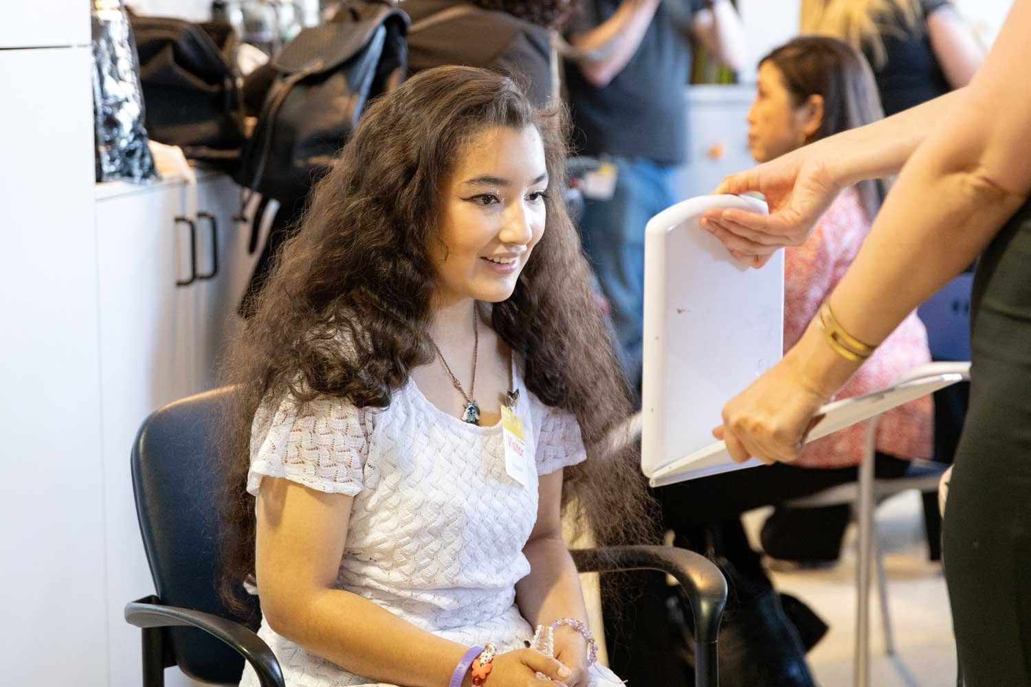 A young teenage girl smiles as she is shown her reflection in a mirror. She has gotten her makeup done for a prom celebration at U C S F Benioff Children's Hospital Oakland.