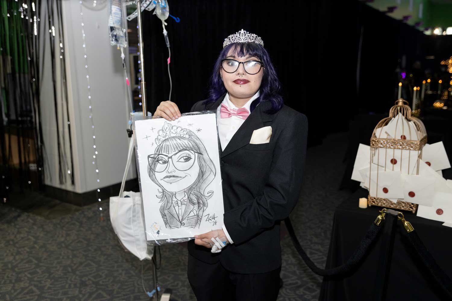 At the U C S F Benioff Children's Hospital Oakland's prom event, a young teenage girl wearing a suit, bow tie, and tiara holds a caricature portrait of herself. Next to her is her IV pole, and prom decorations are in the background.