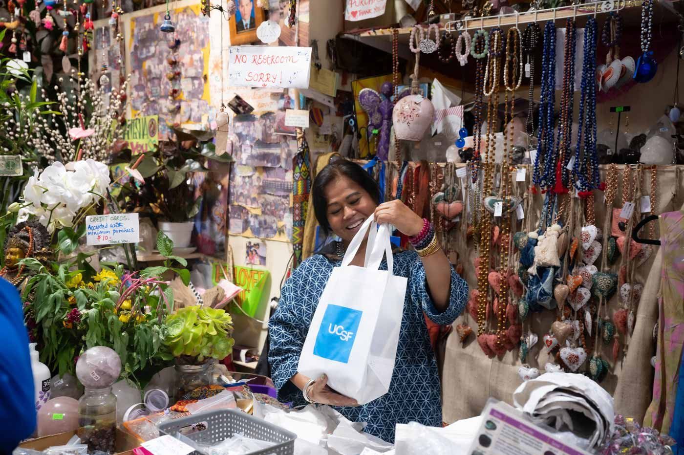 A shop owner on Haight Ashbury stands in her shop and holds a tote bag with the U C S F logo.