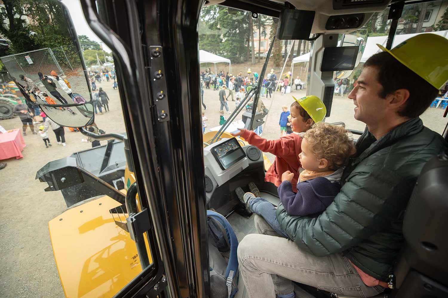 Two children and their dad sit in the driver's seat of a front loader during a Touch a Truck event at Parnassus Heights.