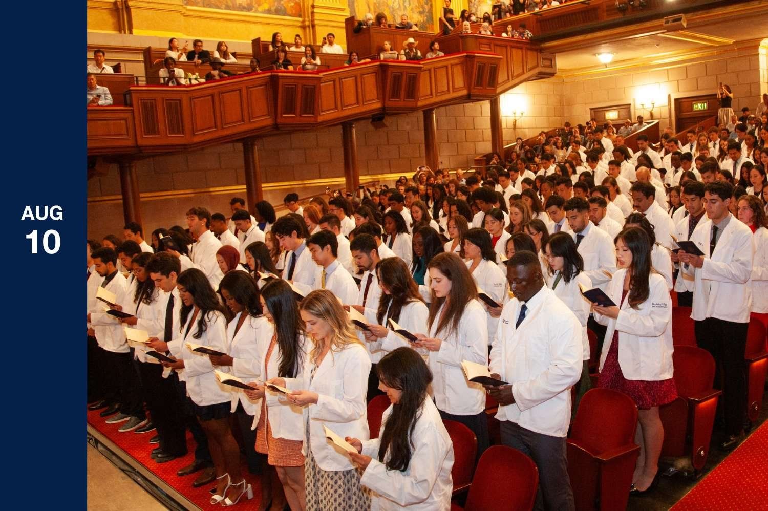 Theater full of students wearing white coats read books in unison.